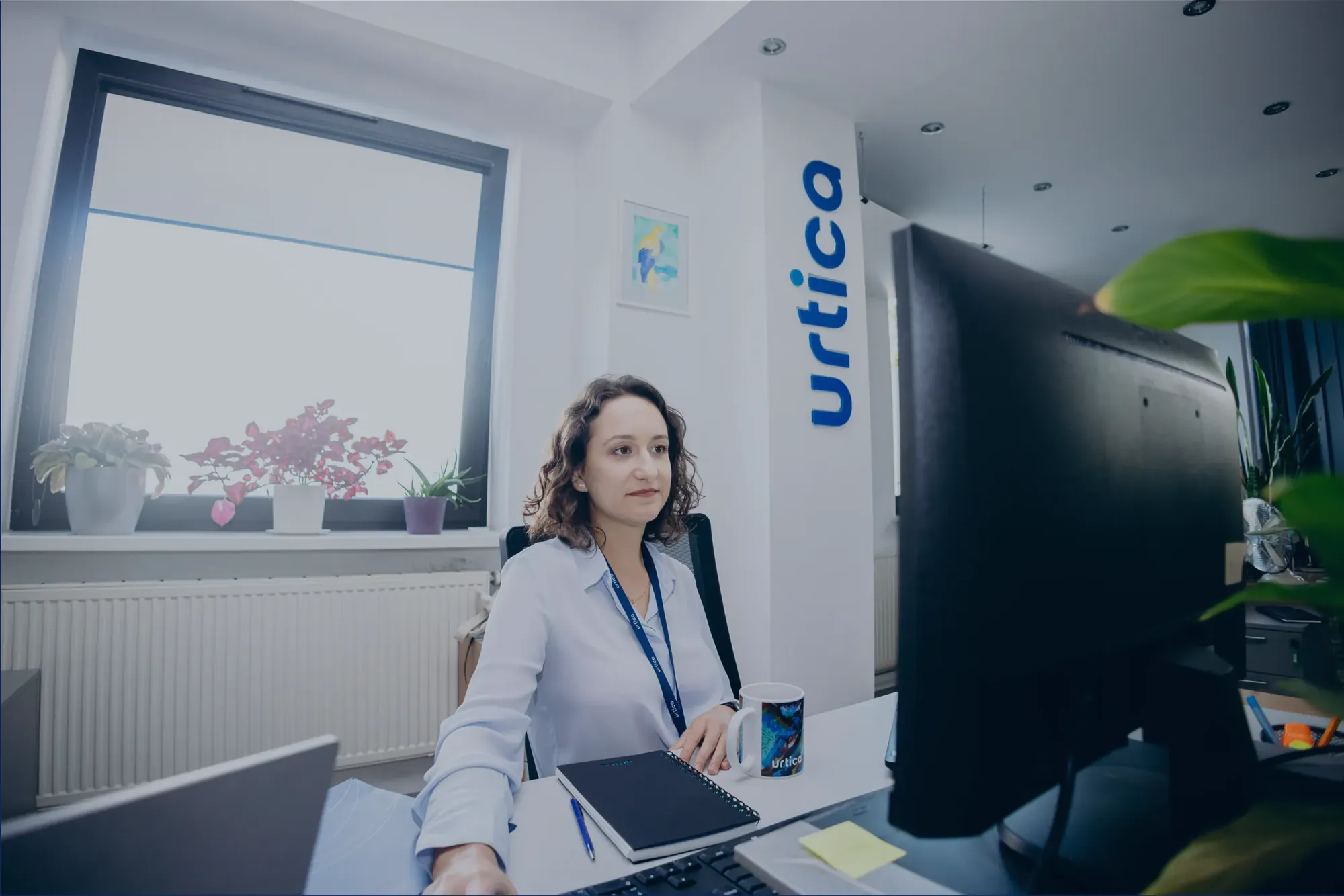 Kobieta przy biurku z notesem i kubkiem, za nią pionowe logo Urtica na ścianie.