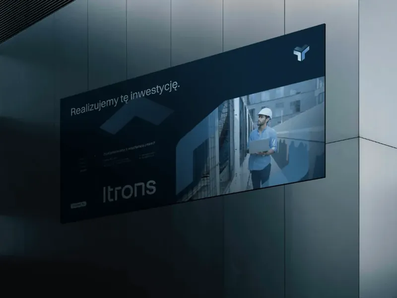 Granatowa tablica Itrons na elewacji z hasłem „Realizujemy tę inwestycję.” i zdjęciem montera w kasku.